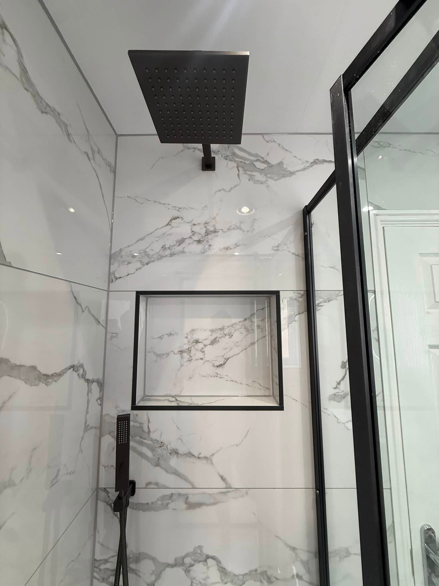 Close-up of the walk-in shower with black rainfall shower, built-in niche and marble-effect wall tiles.