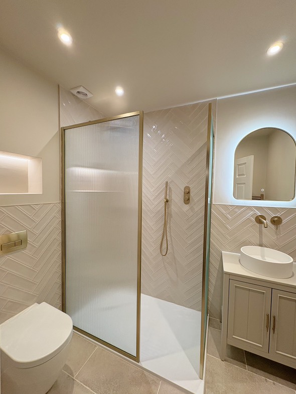 A stylish shower room featuring an Amien brushed gold framed fluted glass screen and Navajo Blanco gloss tiles laid in a herringbone pattern, finished with Elora brushed gold brassware.