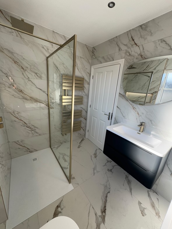 A modern walk-in shower with an Amien brushed gold framed screen and Peoria white slate-effect tray, paired with Stockholm Glenza marble-effect tiles and an Ellis matt black wall-hung vanity.
