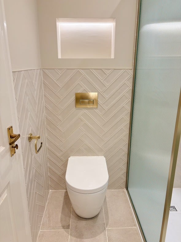 An Oscar back-to-wall toilet featuring a brushed gold flush plate and matching gold toilet roll holder, set against Navajo Blanco gloss tiles in a herringbone pattern with a recessed wall niche.
