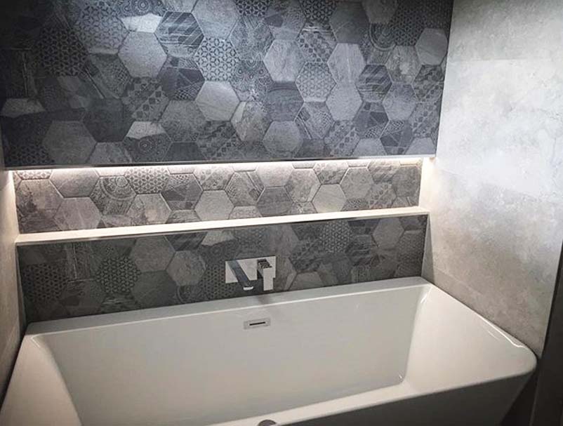 Grey bathroom with patterned hexagon wall tiles, illuminated niche shelf, white freestanding bath and chrome wall-mounted bath filler tap.