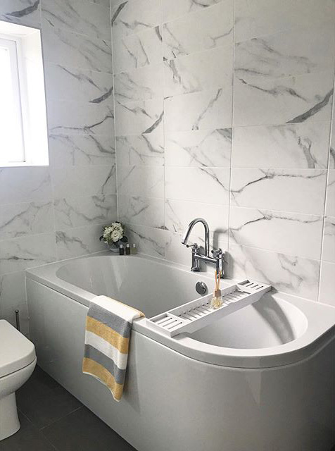 A modern white shower bath featuring a chrome deck-mounted mixer tap and a white wooden bath bridge, set against large-format white marble effect wall tiles with grey veining