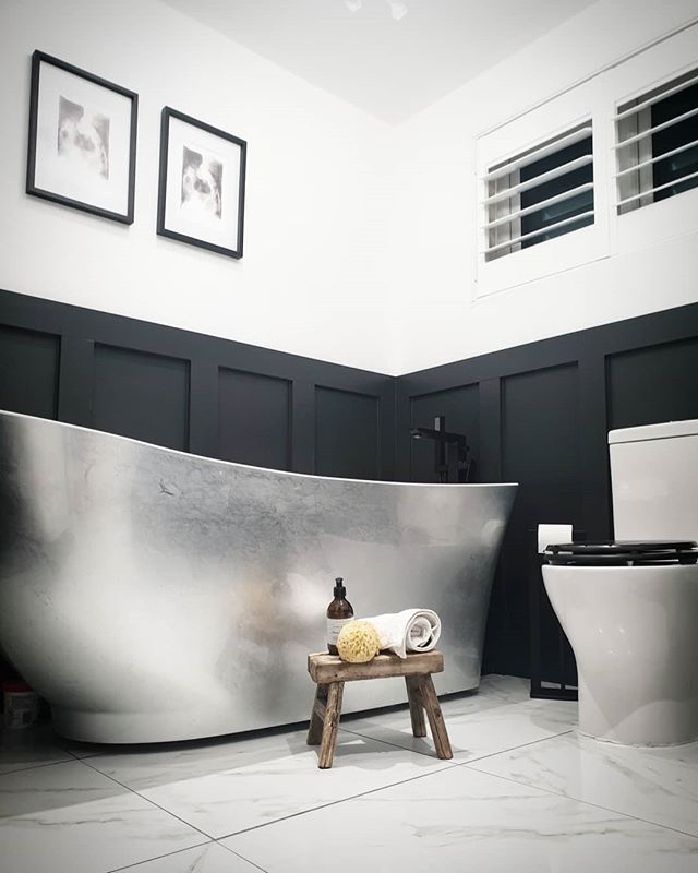A striking metallic silver-finished freestanding bateau bath set against matte black wood wall panelling and white marble-effect floor tiles.
