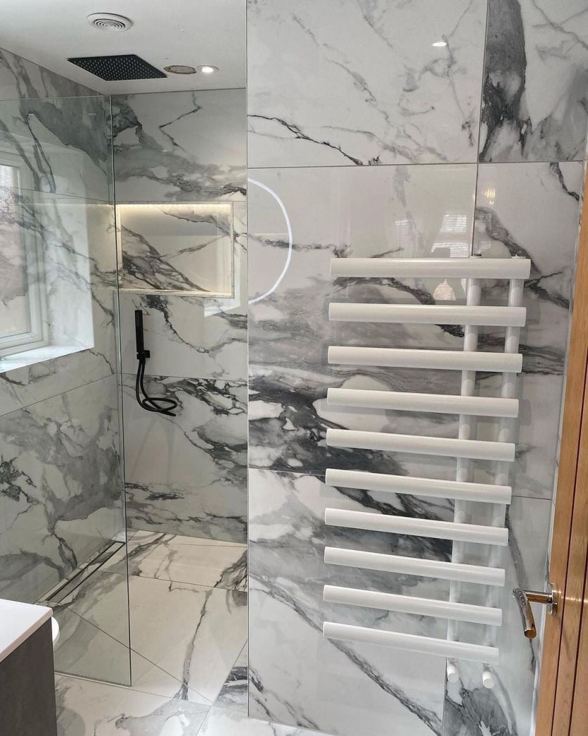 A contemporary walk-in shower featuring large-format white and grey marble effect wall tiles, a recessed wall niche with integrated lighting, and a sleek matte black rainfall shower head and handset.