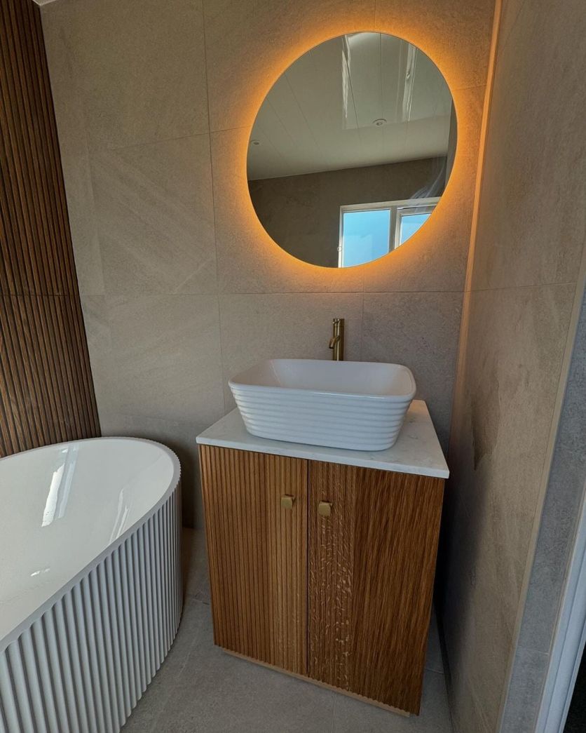 A harmonious spa-style bathroom featuring a Calgary ribbed rectangle basin on a fluted wood vanity, a Felix backlit LED mirror, and an Olympia ribbed freestanding bath against stone-effect and slatted wood tiles.