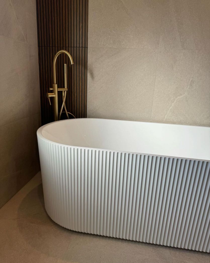 A serene bathroom corner featuring the Olympia white ribbed freestanding bath and Elora brushed gold floor-standing mixer, set against Ottoman slatted wood and Cave Arena stone-effect tiles.