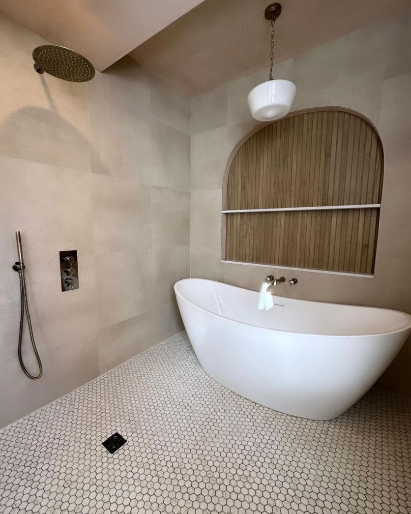 A serene Japandi-style bathroom featuring the Lambi freestanding white bath set against a Dune 4D wood panel tile arched feature wall, with white hexagon mosaic flooring and sandstone-effect wall tiles.