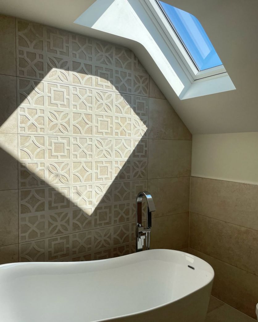 A bright loft bathroom featuring a Tadeo white freestanding acrylic bath paired with a Tear chrome freestanding mixer tap against a feature wall of Reno Avra Marfil textured geometric tiles.