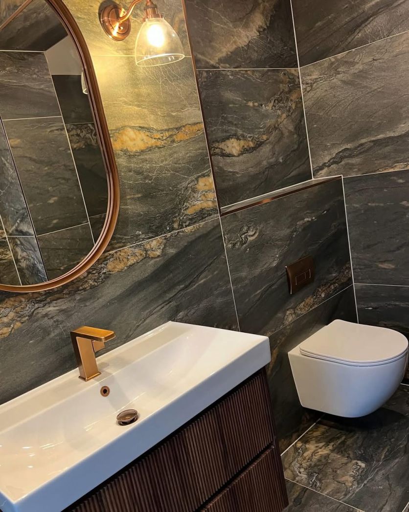 A moody, luxurious bathroom featuring Duomo dark grey marble-effect porcelain tiles, a dark wood ribbed vanity with a Flint brushed bronze tap, and an Oscar wall-hung toilet with a matching bronze flush plate.