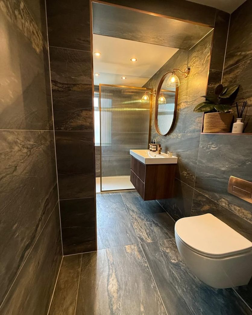 A wide view of a moody industrial bathroom featuring Duomo dark grey marble-effect tiles on walls and floor, an Amien brushed bronze framed fluted glass shower screen, and a wall-hung vanity with bronze accents.