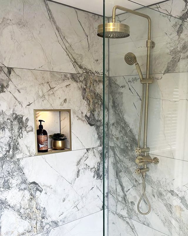 A luxurious walk-in shower featuring large-format white and grey marble effect wall tiles. The space is elevated by a sophisticated brushed gold thermostatic rainfall shower kit