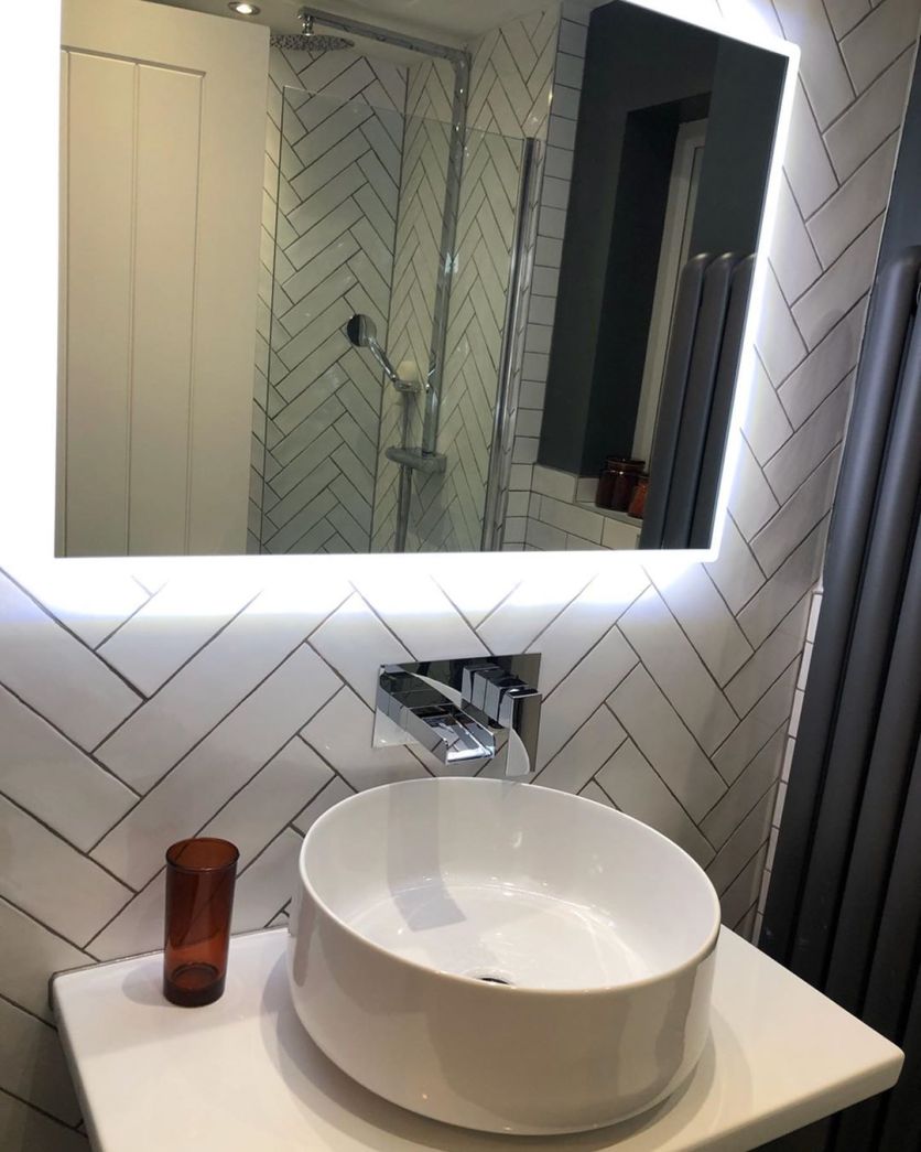 A white countertop vessel basin with a chrome wall-mounted waterfall tap, set against white herringbone tiles and an illuminated LED mirror.