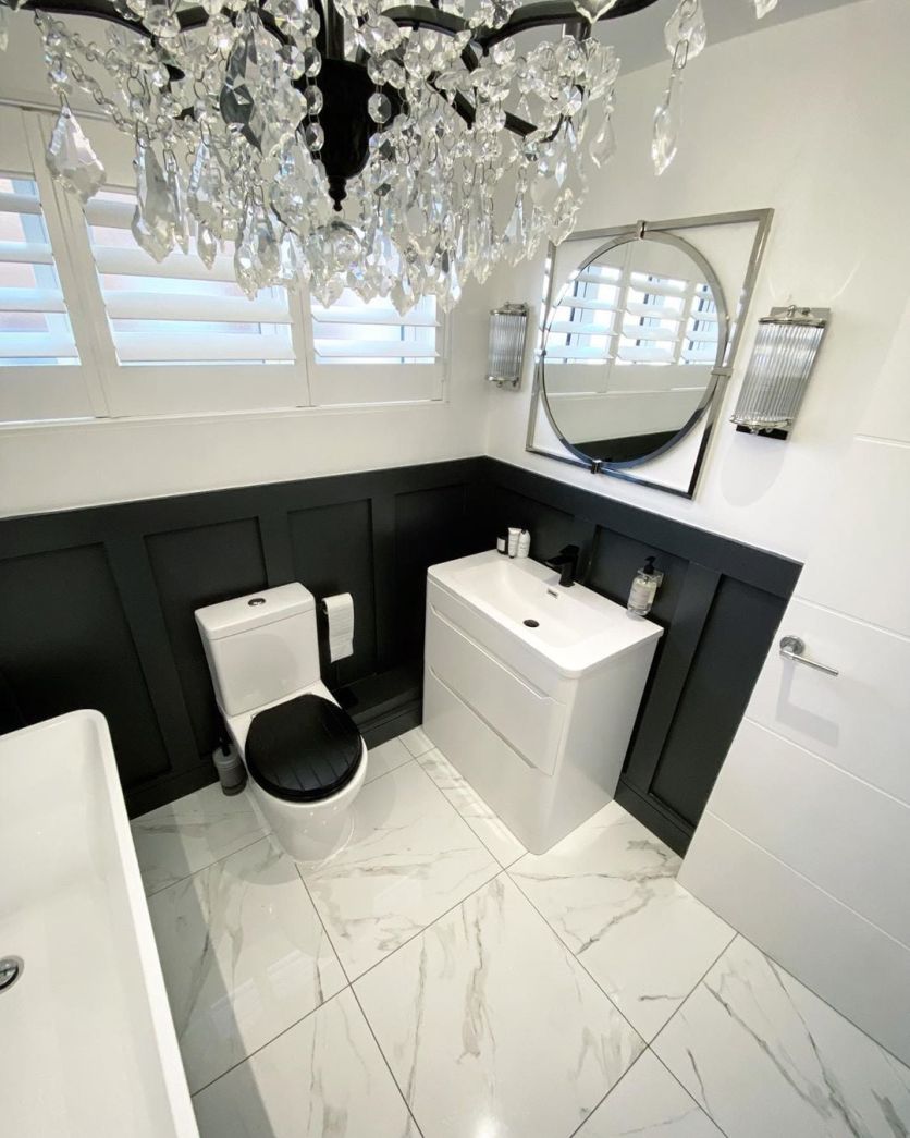 A luxurious traditional bathroom featuring dramatic black wall panelling, white marble-effect floor tiles, and a white gloss vanity unit. The space is finished with a stunning crystal chandelier and a circular silver-framed mirror.