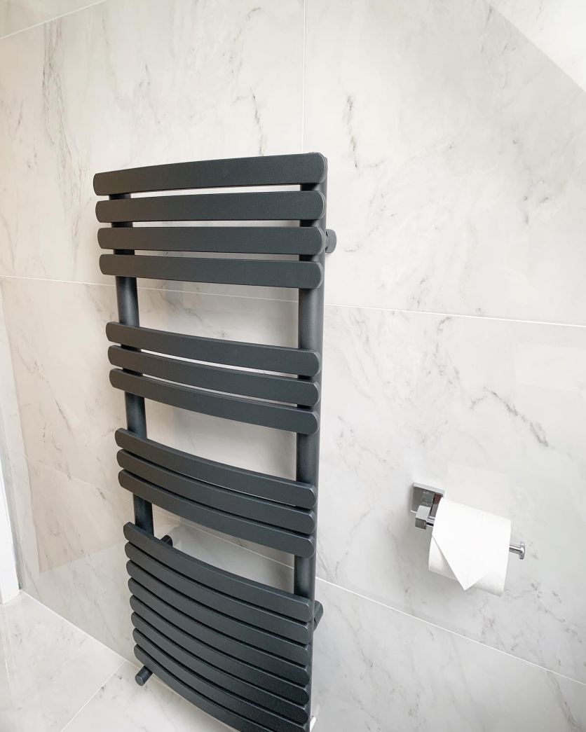 A designer anthracite grey heated towel radiator with flat panels mounted on white marble-effect wall tiles next to a square chrome toilet roll holder.