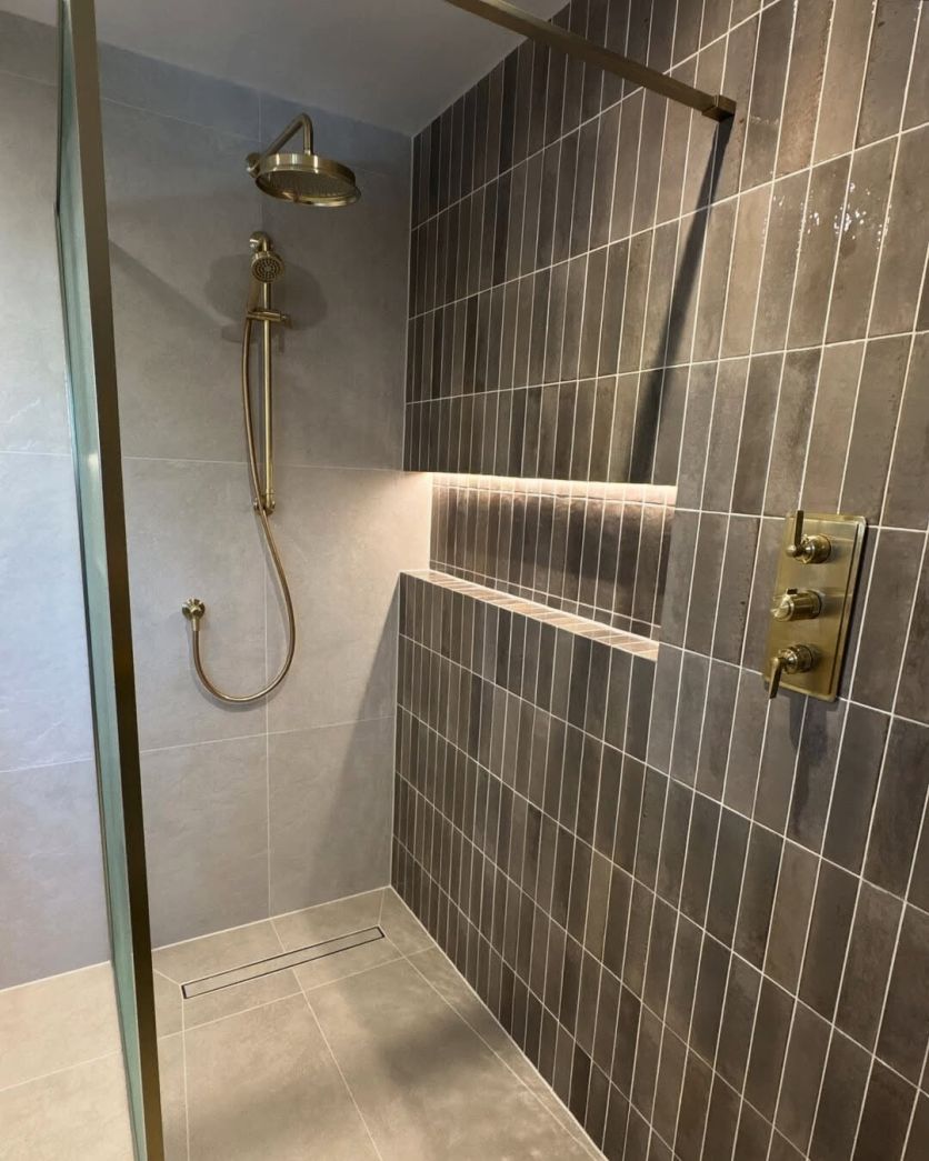 A modern walk-in shower featuring dark grey vertical wall tile and a brushed gold shower head and valves.
