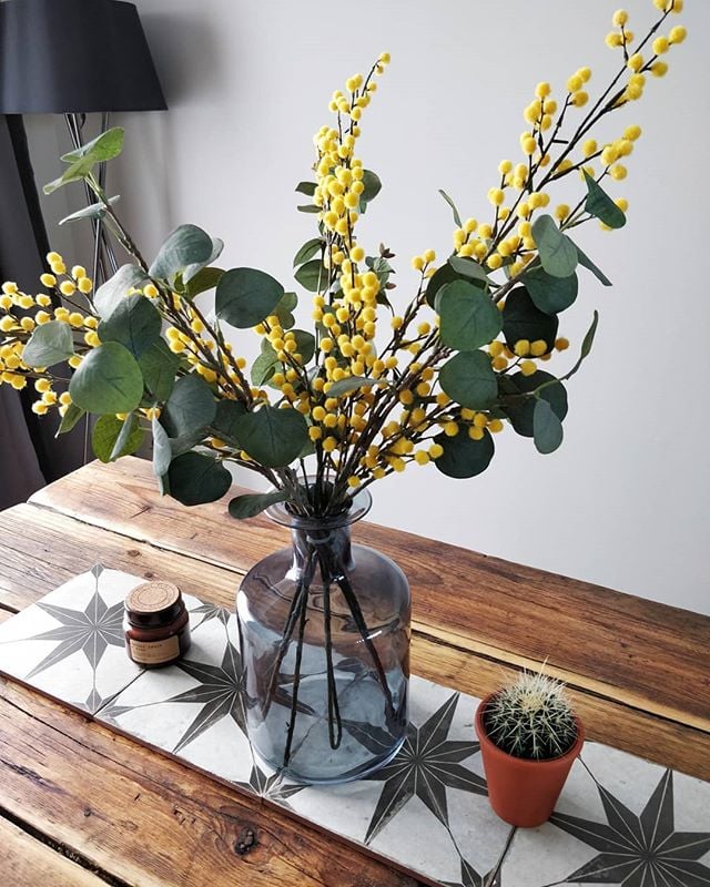 Grey and white star patterned tiles inlaid on a rustic wooden surface next to a glass vase with yellow mimosa flowers and greenery.