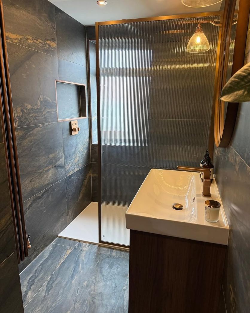 A luxurious, moody bathroom featuring Duomo dark grey marble-effect porcelain tiles, an Amien brushed bronze framed fluted glass shower screen, and a dark wood vanity with Flint brushed bronze brassware.