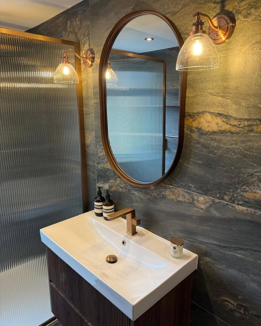 A luxurious moody bathroom featuring Duomo dark grey marble-effect tiles, an Amien brushed bronze framed fluted glass shower screen, and a dark wood ribbed vanity with a Flint brushed bronze tap.