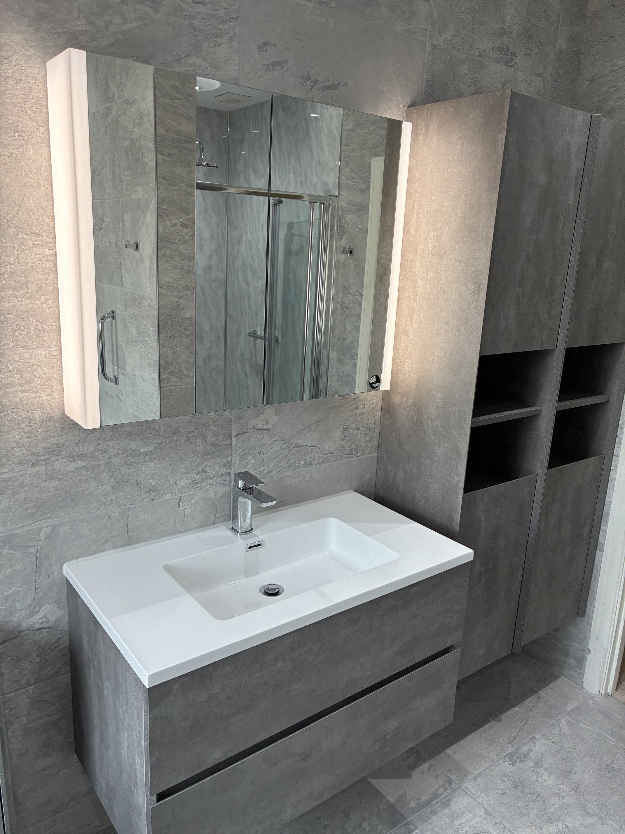 Completed bathroom vanity area with grey furniture, white basin, illuminated mirror and fitted storage.