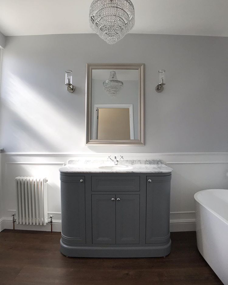 Traditional bathroom with a grey curved vanity, marble top, chrome tap and chandelier above dark wood flooring.