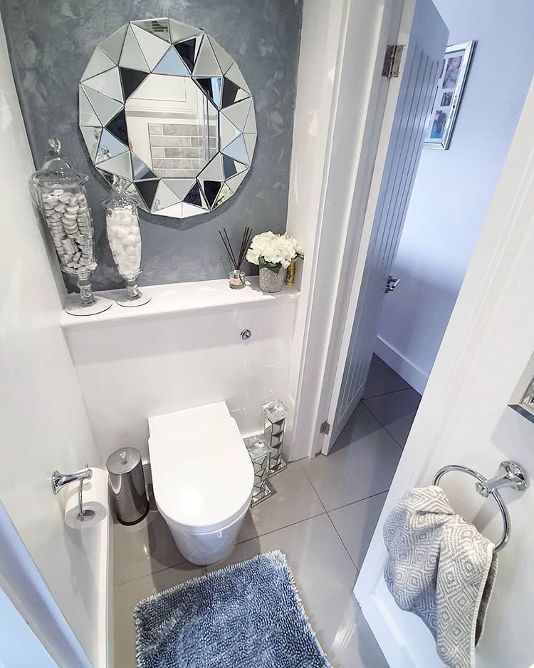 A stylish cloakroom featuring a modern white back-to-wall toilet with a hidden cistern, set against a textured grey feature wall. Above the toilet hangs a striking, large faceted geometric mirror, complemented by grey floor tiles and chrome accessories.