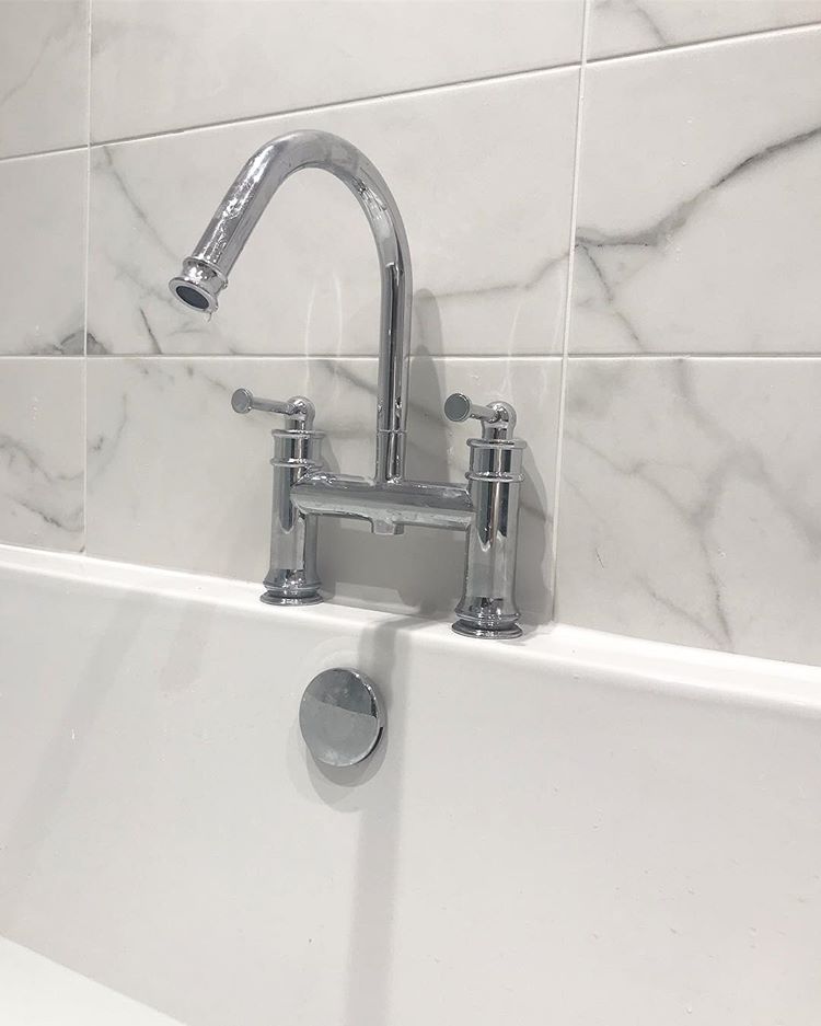 Traditional chrome bridge bath mixer tap with lever handles mounted on a white bath against grey-veined marble effect wall tiles.