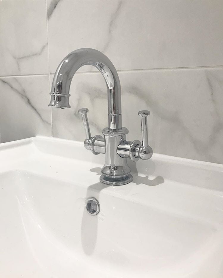 A traditional chrome mono basin mixer tap with elegant lever handles, mounted on a white ceramic sink against marble-veined wall tiles.