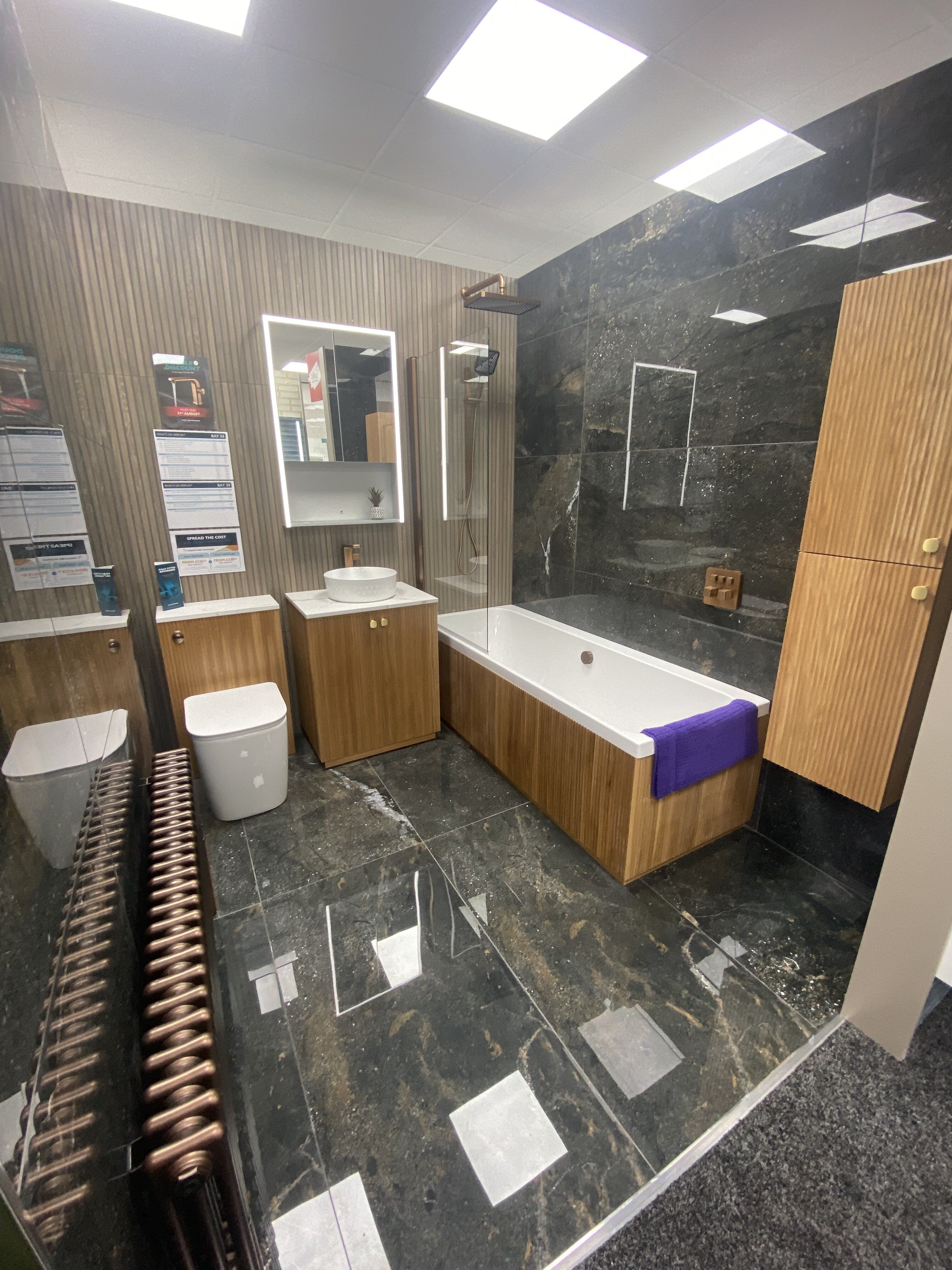 New Newbury bathroom display featuring warm wood furniture, dark wall tiles and illuminated mirrors.