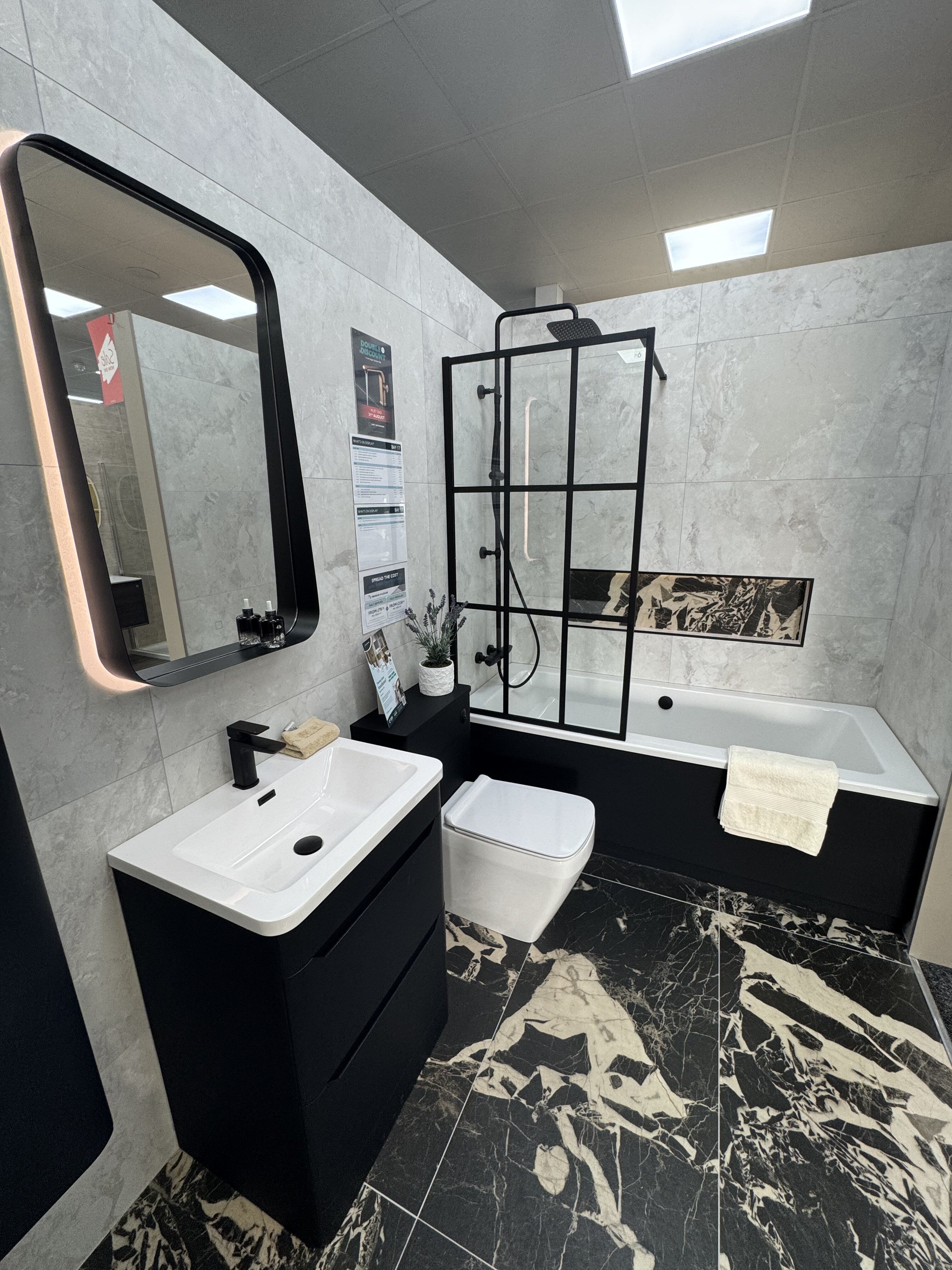 Full view of the modern bathroom display featuring black furniture, marble-effect tiles, bath and framed shower screen.