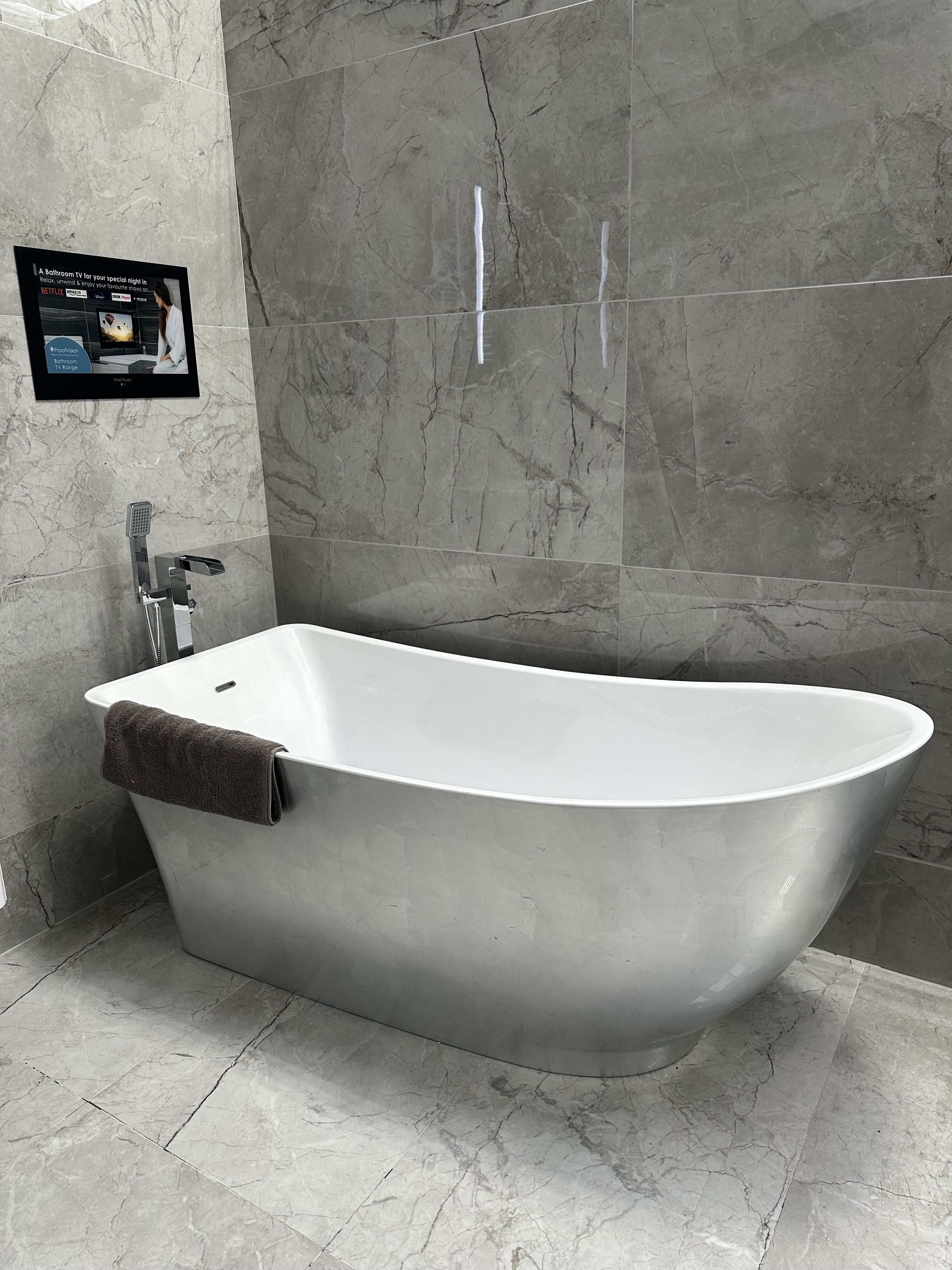 Essence silver freestanding bath displayed against grey marble-effect tiles with a built-in wall-mounted TV at Easy Bathrooms Watford.