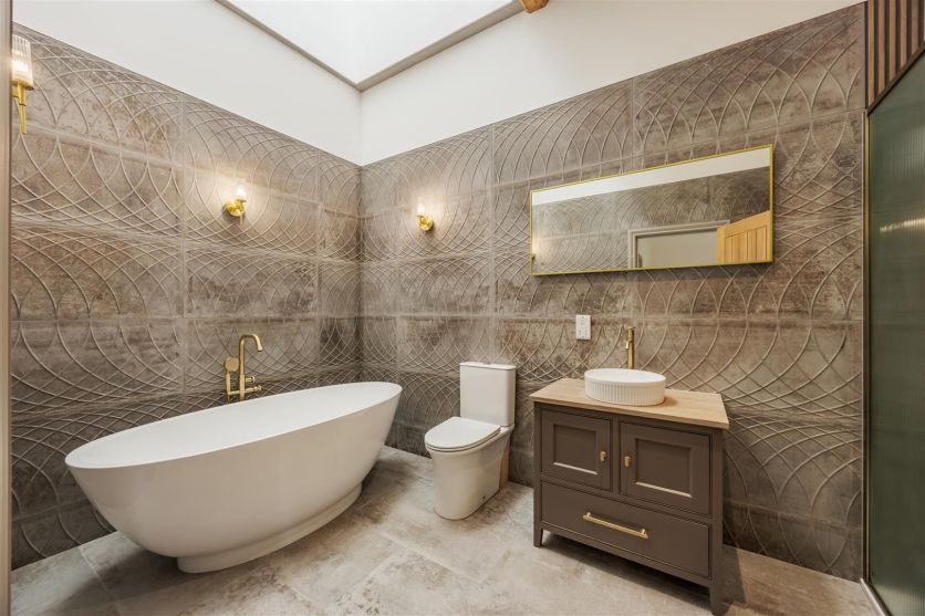 A luxury bathroom featuring Fontana Ceniza textured decor wall tiles and a white freestanding bath with an Urban brushed gold mixer tap, complemented by a grey vanity unit with a ribbed countertop basin and a large Rione gold backlit mirror.