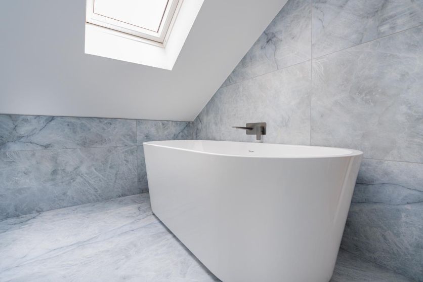 A Noah white freestanding acrylic bath paired with a wall-mounted gunmetal tap, set against Kendrick Sky blue marble-effect porcelain tiles under a vaulted ceiling with a skylight.