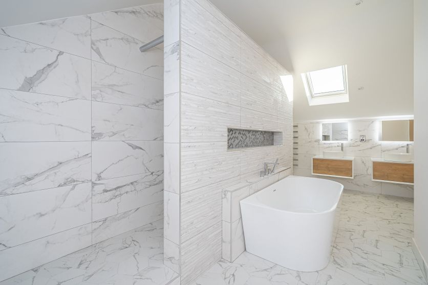 A bright, spacious loft bathroom featuring white marbleffect wall tiles and matching hexagon floor tiles. A white D-shaped back-to-wall bath, complemented by twin floating oak vanity units with white countertop basins.