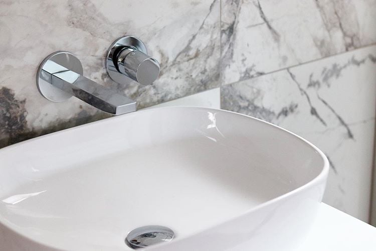 A white oval countertop vessel basin featuring sleek wall-mounted chrome mixer taps set against grey and white marble-effect porcelain wall tiles.