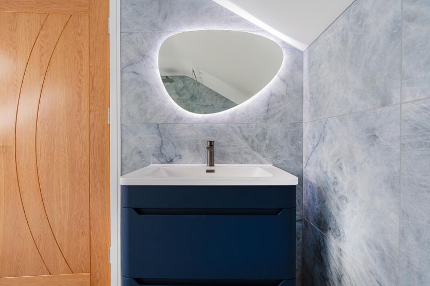 A modern vanity area featuring an Ellis royal blue floor-standing unit with a white basin and gunmetal tap, set against Kendrick Sky blue marble-effect porcelain tiles and an Ivy backlit pebble mirror.
