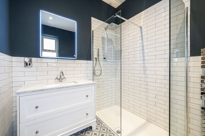 A traditional white vanity unit with a marble top and chrome taps, positioned next to a modern walk-in shower with white metro tiling, set against a bold navy blue wall and patterned Victorian-style floor tiles.