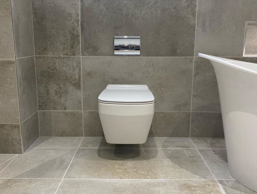 A minimalist wall-hung white toilet with a sleek chrome dual-flush plate, mounted against large-format grey stone effect wall tiles.
