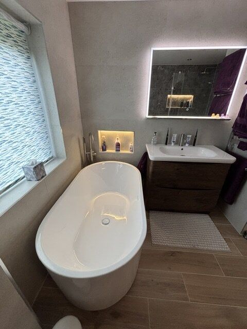 Completed bathroom with a freestanding bath, illuminated wall niche and warm wood-effect flooring.