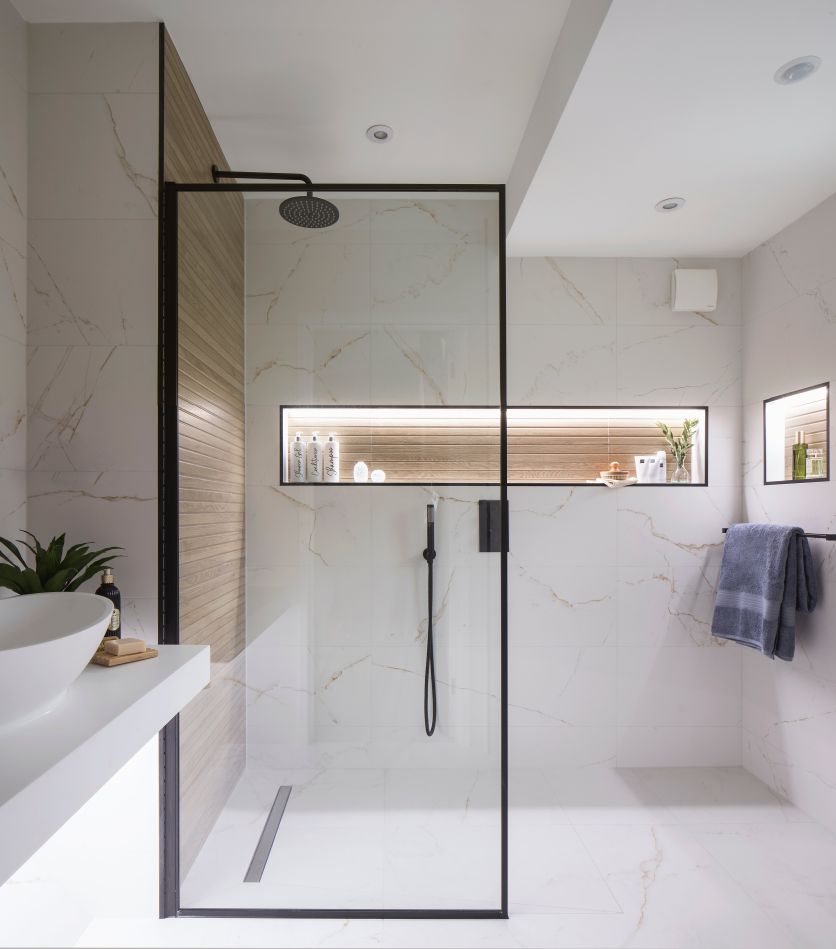 A luxury wet room featuring a black rainfall shower and framed glass screen, set against white gold-veined marble tiles. A long, illuminated recessed shelf with wood-effect backing adds a warm glow, complemented by a sleek white countertop basin.