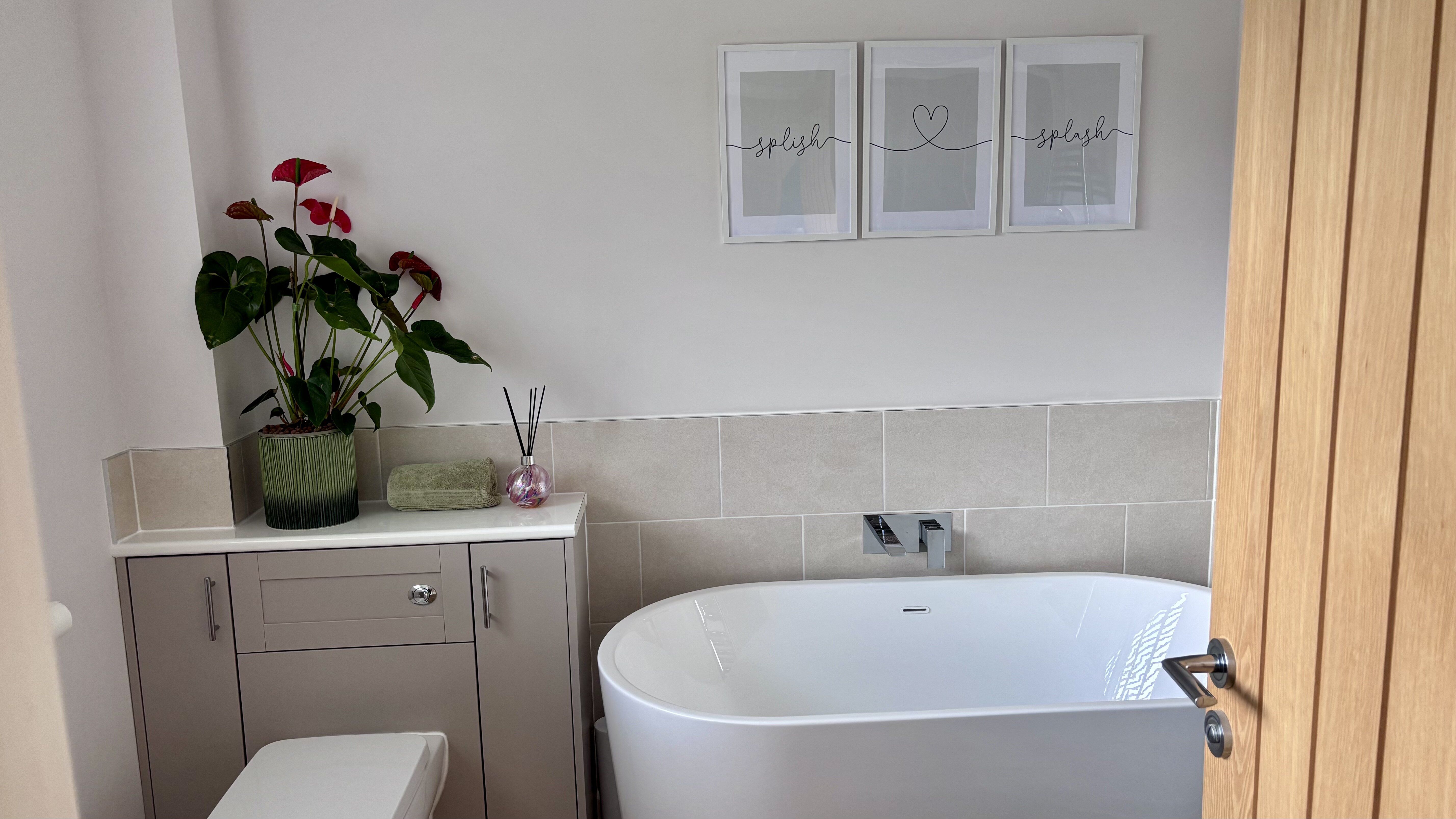Freestanding white bath with neutral furniture and soft marble-effect wall tiles.