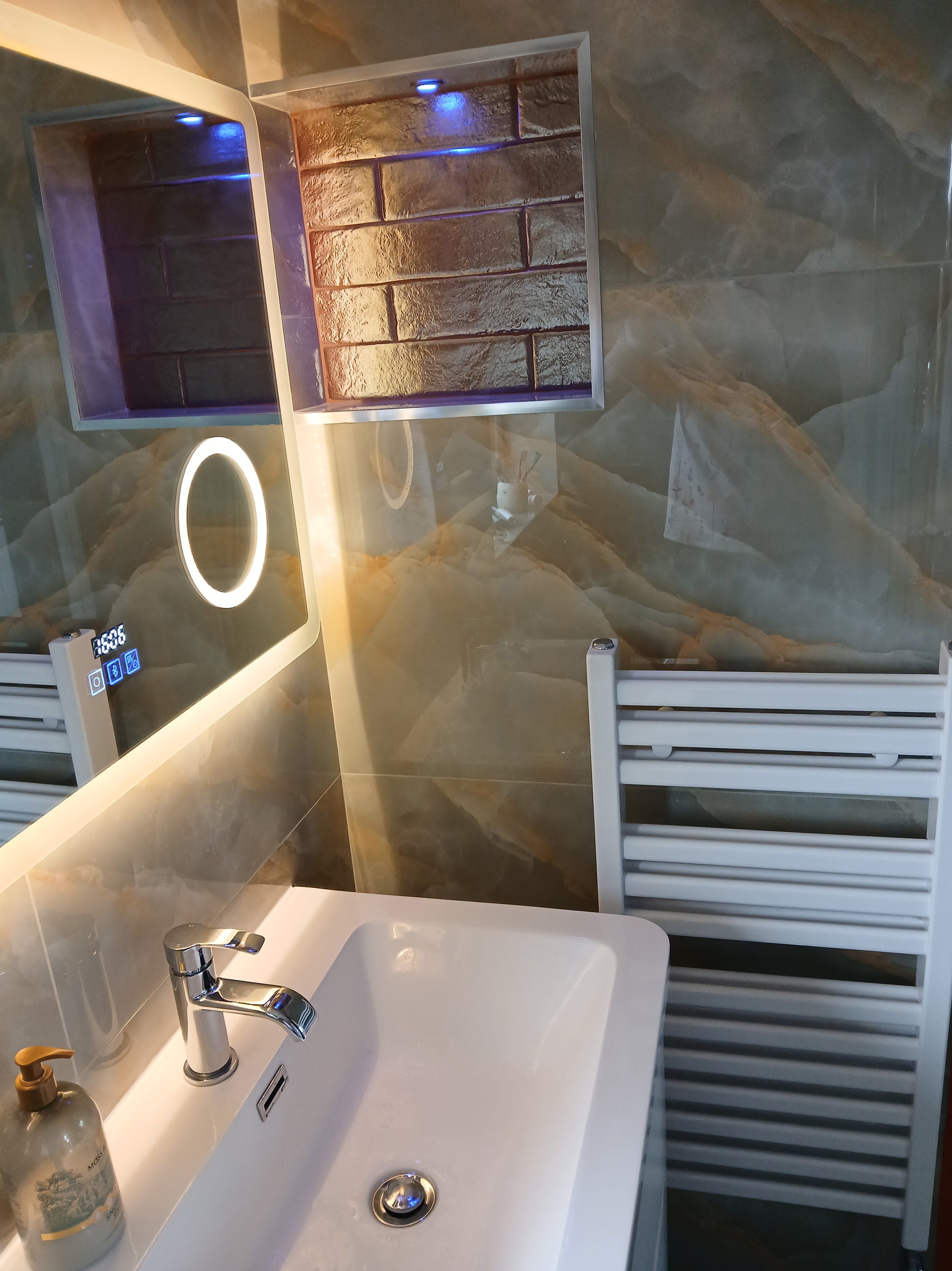 Bathroom basin with chrome tap, illuminated mirror, display niche and white heated towel rail against marble-effect tiles.