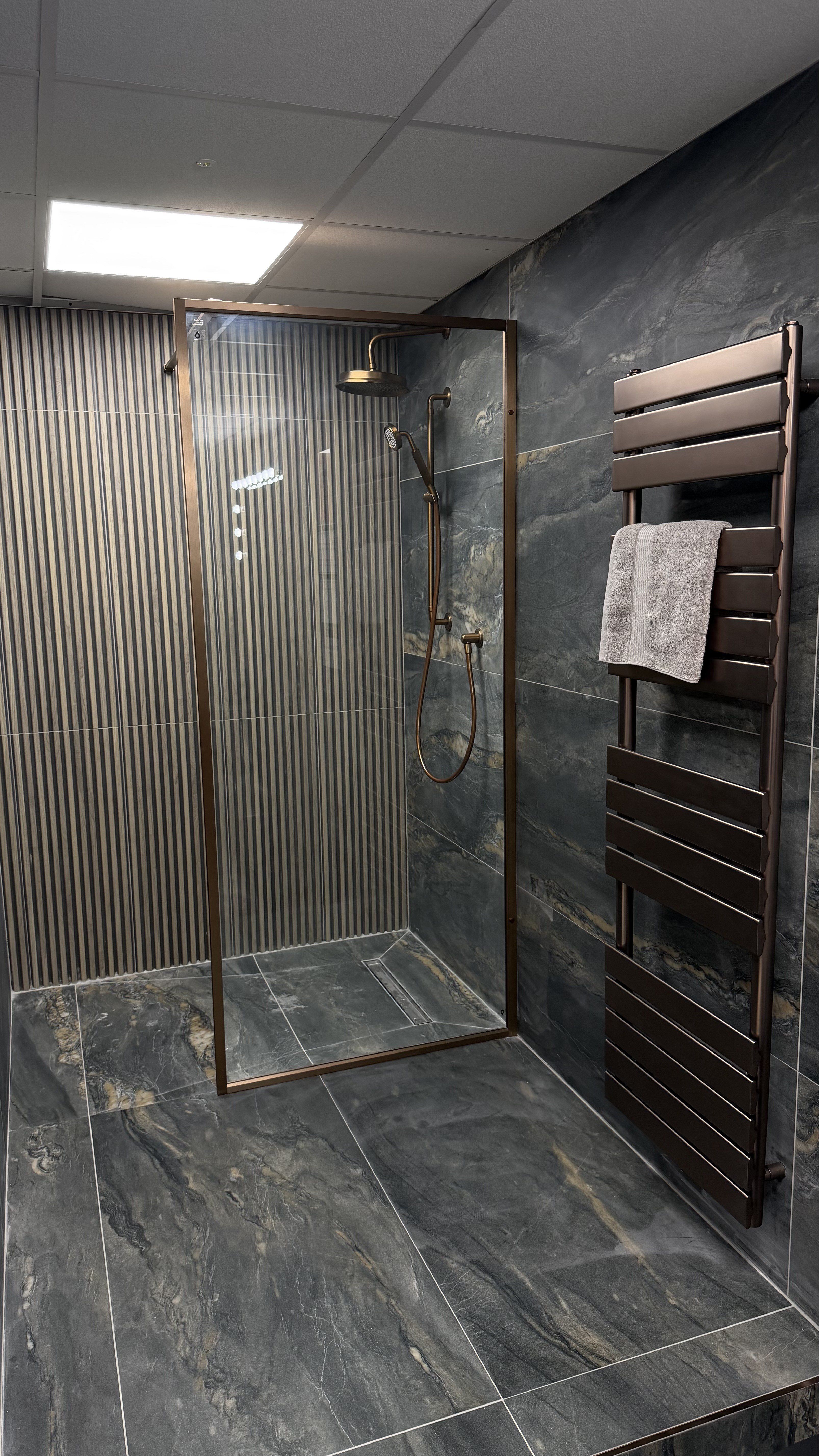 Walk-in shower display with brushed bronze frame, dark stone-effect tiles and coordinating radiator.