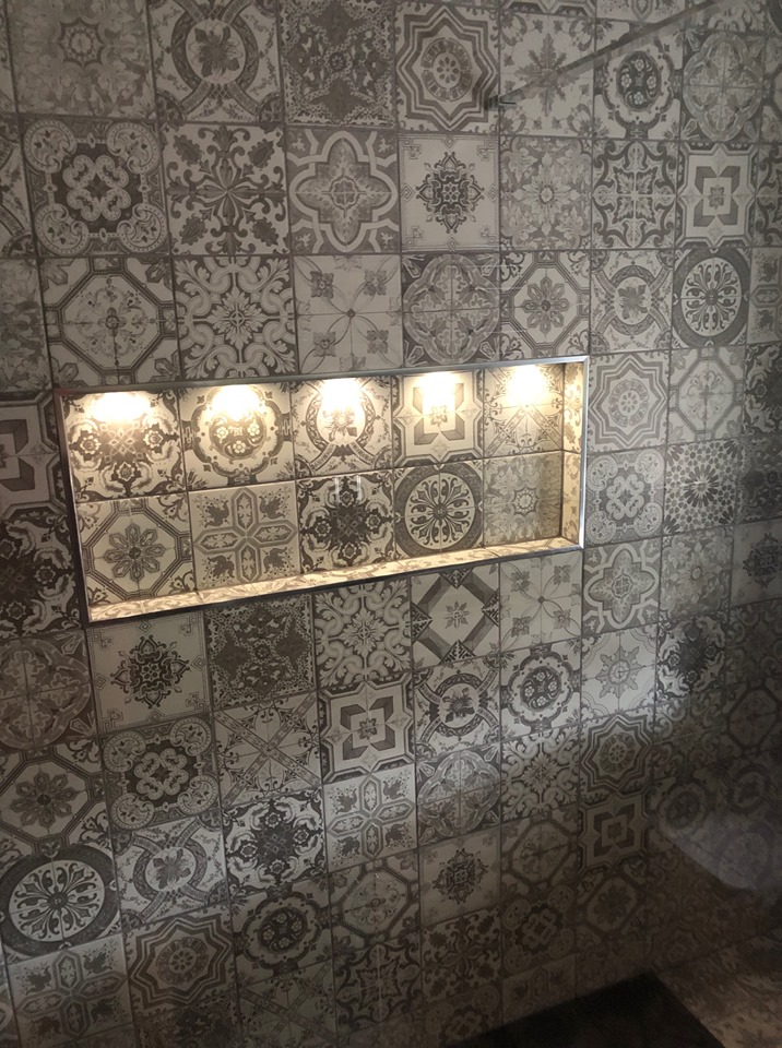Intricate grey and white patterned feature wall tiles arranged in a patchwork style, featuring a recessed shower niche with integrated warm LED spotlights and a sleek chrome trim.