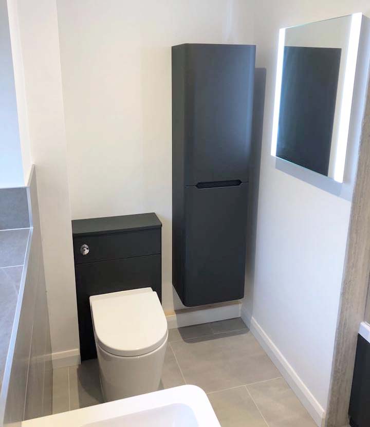 A compact modern bathroom featuring a graphite grey back-to-wall toilet unit and a matching wall-hung tall storage cabinet, set against neutral walls with a slimline LED illuminated mirror.