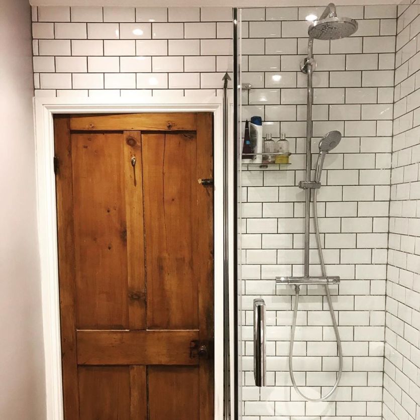 A chrome thermostatic rainfall shower with a handheld attachment, set against white metro tiles with dark grout, beside a rustic wooden period door.