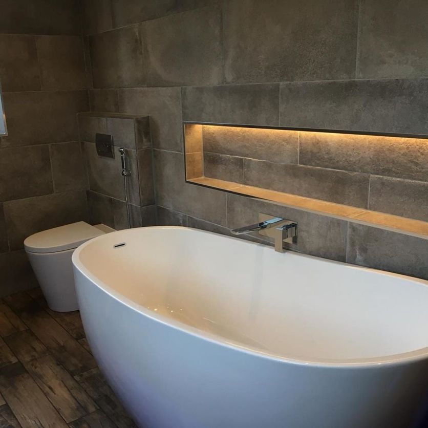 A sleek white oval freestanding bath with a wall-mounted chrome tap, set against large-format stone-effect wall tiles featuring an illuminated recessed shelving niche and matching wood-effect flooring.