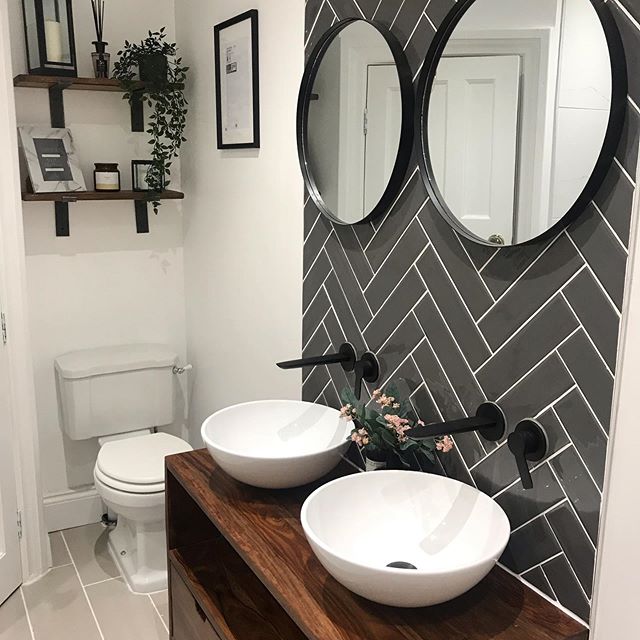 Modern bathroom with grey herringbone tile feature wall, twin white countertop basins, black wall-mounted taps and wooden vanity unit.