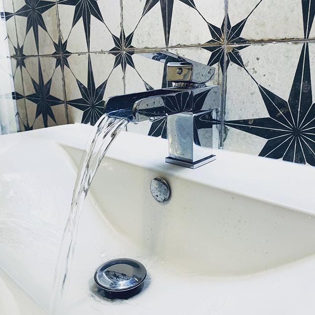 A close-up of a contemporary chrome waterfall basin mixer tap flowing into a white ceramic sink, set against a striking monochrome star-patterned feature tile wall.