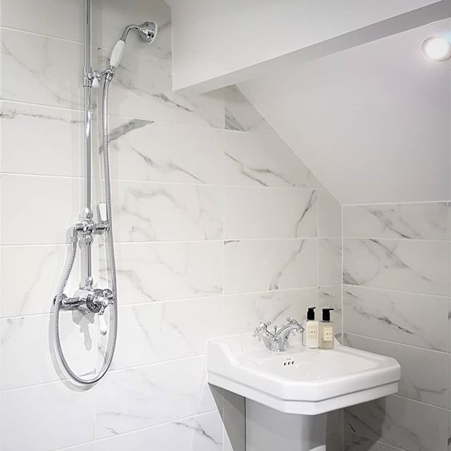 A classic chrome exposed thermostatic shower valve with a white ceramic handle, mounted on large-format white marble effect wall tiles next to a traditional white pedestal washbasin.
