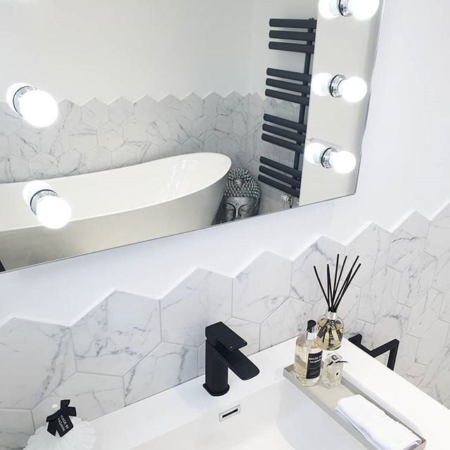 A modern matte black basin tap and white sink set against white marble-effect hexagon tiles, reflected in a large illuminated Hollywood-style vanity mirror.